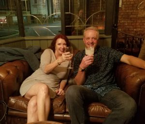 Pam and Dave with drinks, sitting on a leather settee.